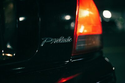 Dramatic close-up of a Honda Prelude emblem and tail light with bokeh background at night.