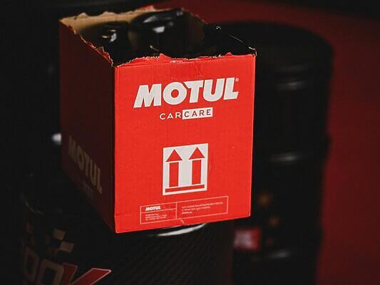 Close-up of Motul car care packaging atop oil barrels in a dark setting.