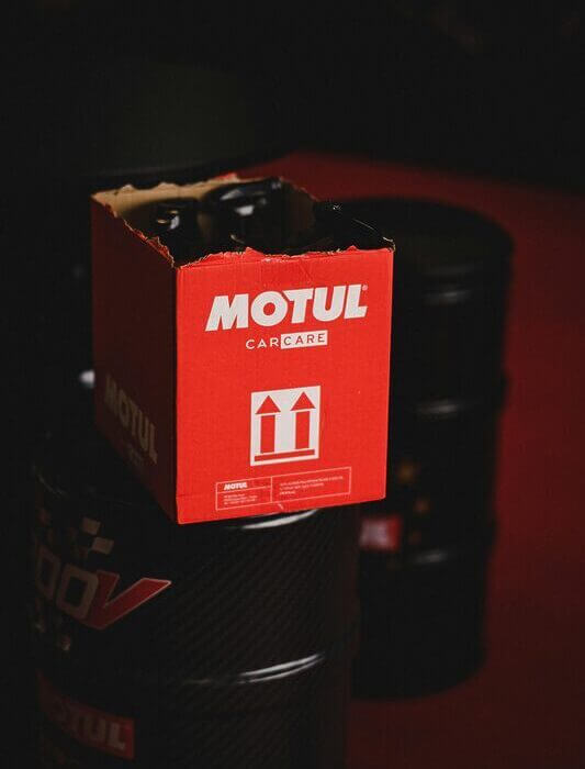 Close-up of Motul car care packaging atop oil barrels in a dark setting.