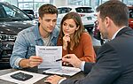 A couple reviews a lease agreement with a salesperson in a car dealership. A calculator and documents are on the table