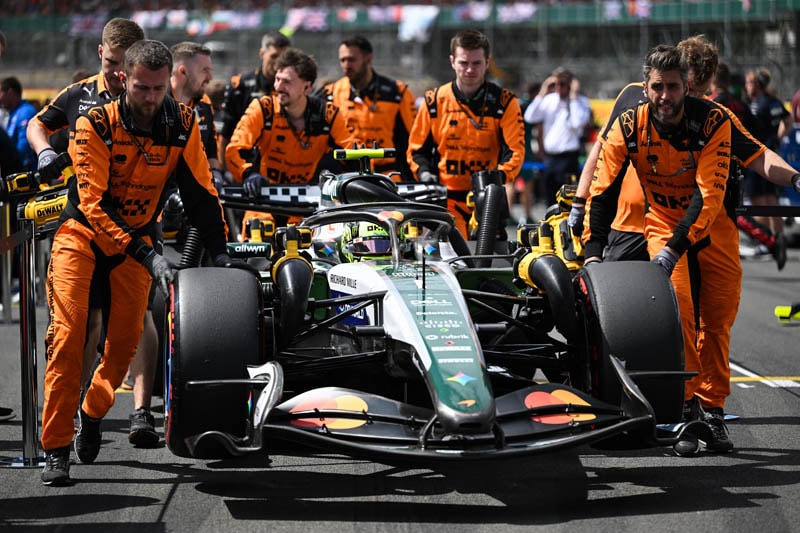 Zak Brown dans le paddock de Silverstone avec la monoplace McLaren équipée d’un moteur Mercedes en arrière-plan.