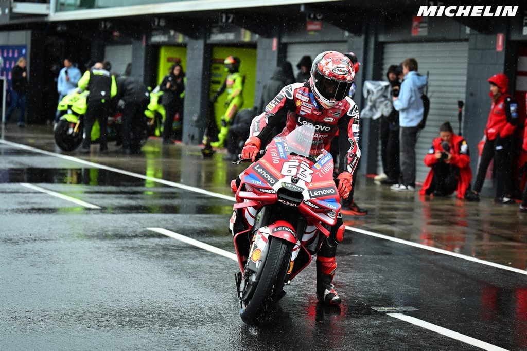 Un pilote en combinaison rouge et noire pousse une Ducati MotoGP rouge dans la voie des stands détrempée. Le numéro 63 et le logo Lenovo sont visibles.
