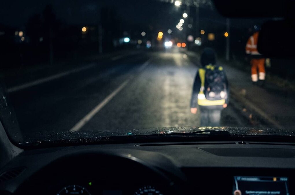 Un piéton portant un gilet réfléchissant devant une voiture dans l'obscurité.