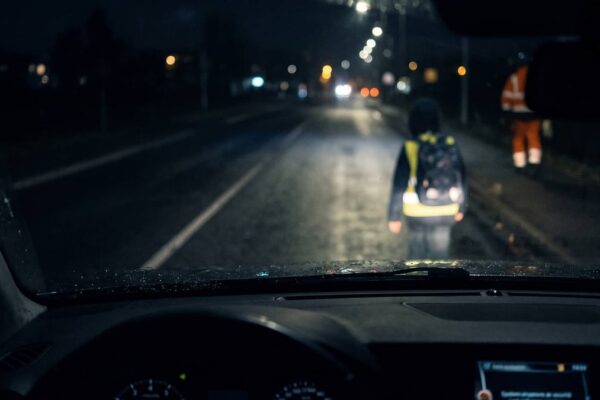 Un piéton portant un gilet réfléchissant devant une voiture dans l'obscurité.