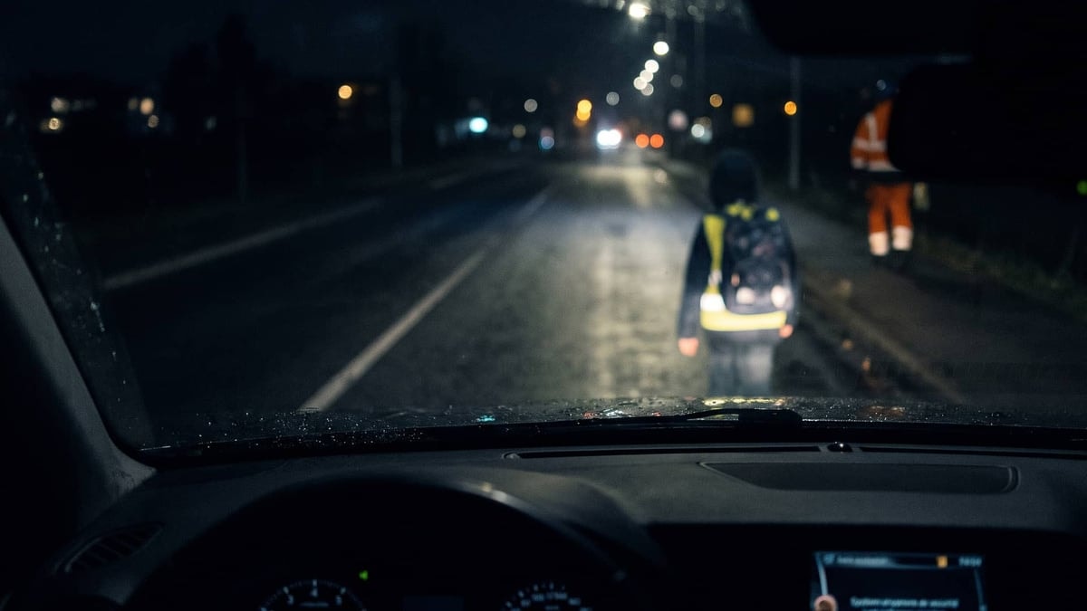 Un piéton portant un gilet réfléchissant devant une voiture dans l'obscurité.